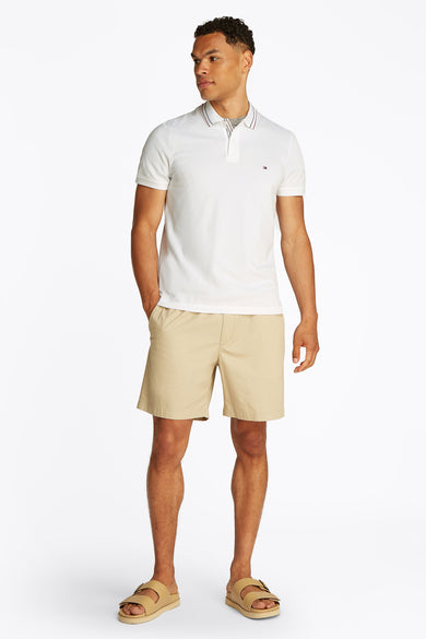 Wit poloshirt met korte mouwen van TOMMY HILFIGER met decoratieve strepen op de kraag, gedragen met een beige korte broek en sandalen.
