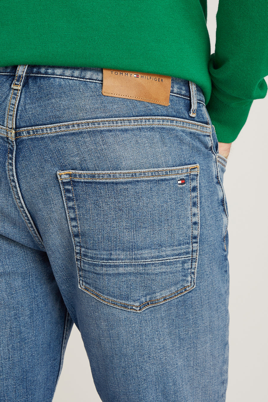 Jean droit en denim bleu clair de Tommy Hilfiger, avec poche arrière ornée du logo, porté avec un pull vert.
