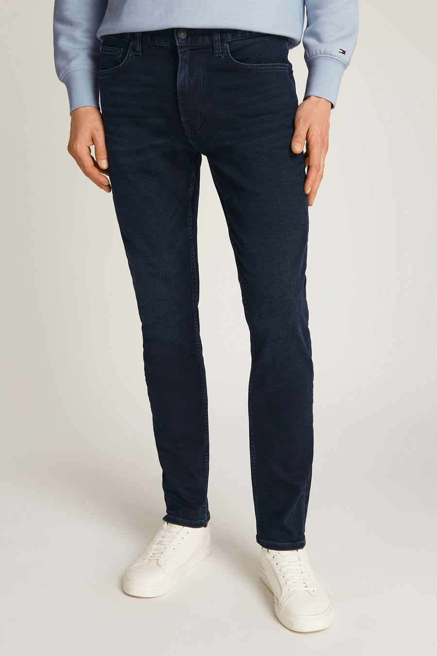 Rechte jeans in zwartblauw denim van TOMMY HILFIGER, gedragen met een lichtblauwe trui en witte sneakers.
