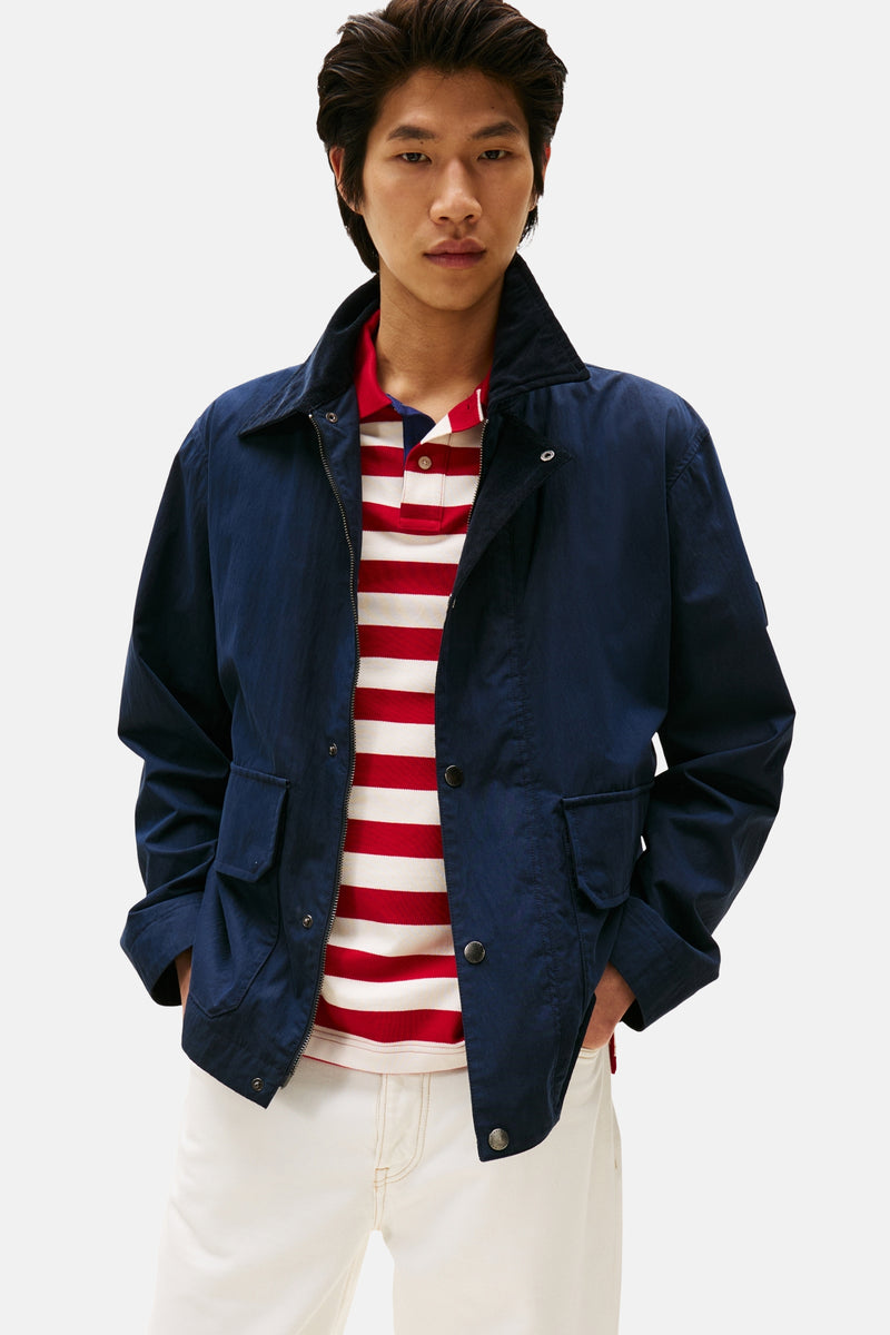 Veste bleue de TOMMY HILFIGER portée sur un polo rayé rouge et blanc avec un pantalon blanc.