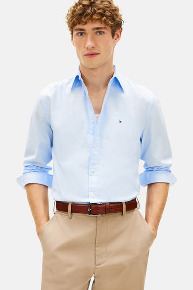 Chemise à manches longues bleu clair de TOMMY HILFIGER, portée ouverte avec un pantalon beige et une ceinture marron.