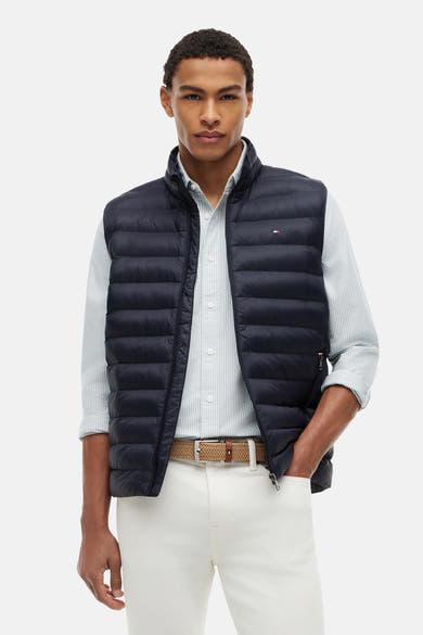 Matblauwe gewatteerde bodywarmer van TOMMY HILFIGER, met klein vlaglogo en gedragen over een gestreept overhemd.