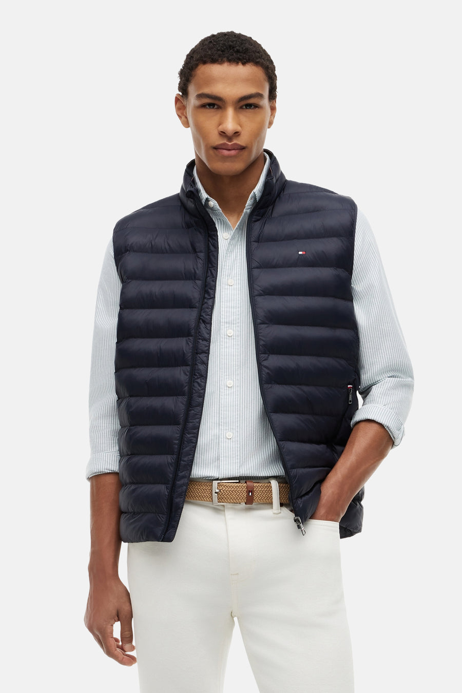 Matblauwe gewatteerde bodywarmer van TOMMY HILFIGER, met klein vlaglogo en gedragen over een gestreept overhemd.