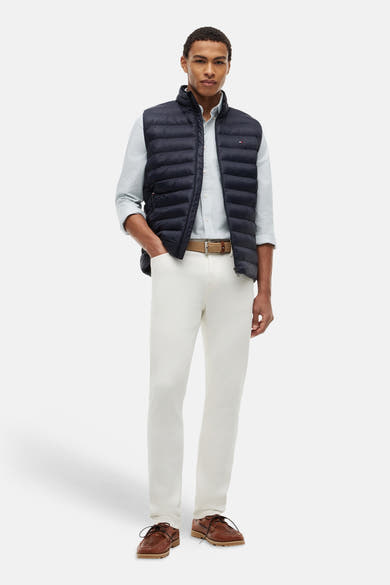 Gewatteerd blauw bodywarmer van TOMMY HILFIGER, gedragen door een man met een gestreept overhemd, een witte broek en bruine schoenen.