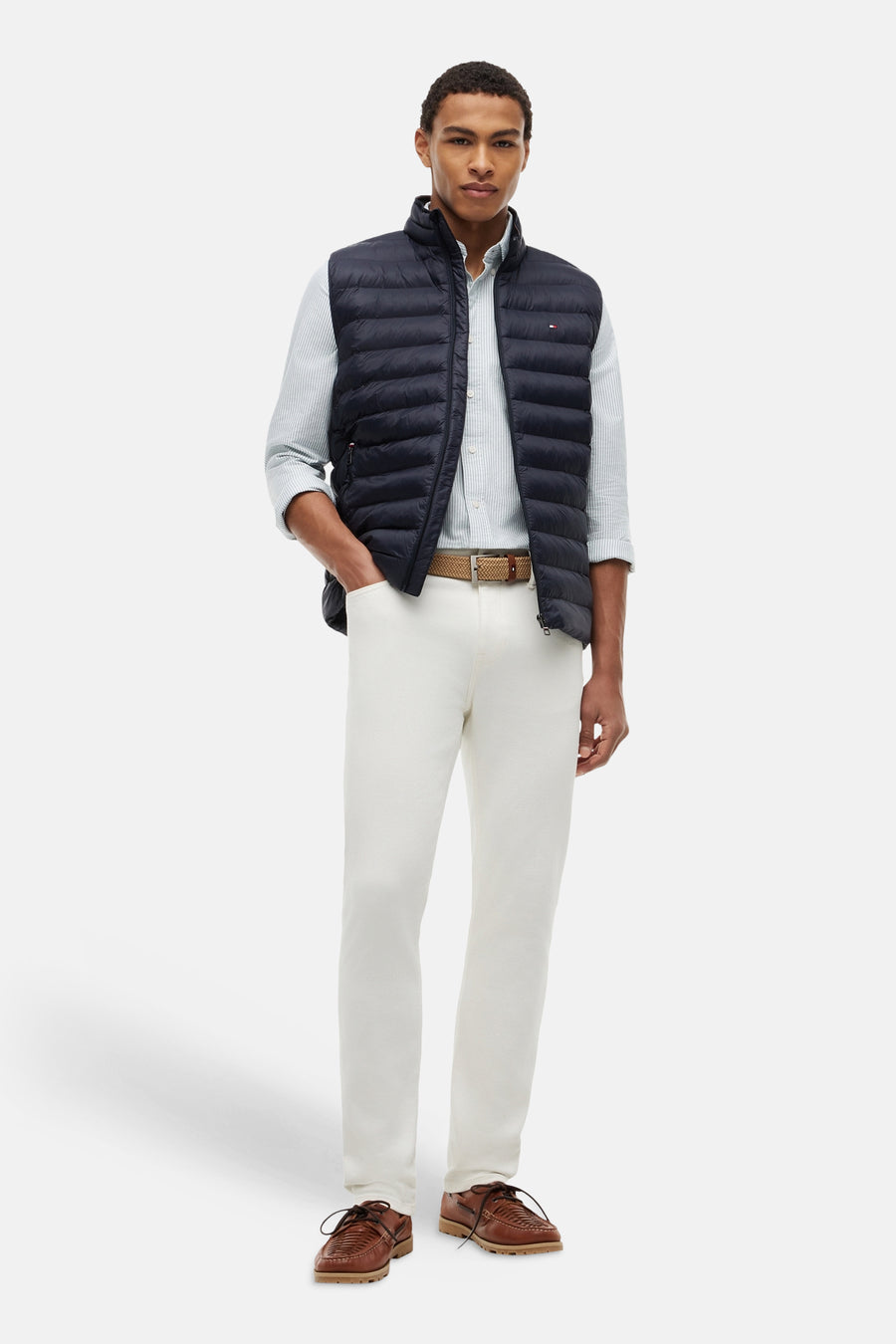 Gewatteerd blauw bodywarmer van TOMMY HILFIGER, gedragen door een man met een gestreept overhemd, een witte broek en bruine schoenen.
