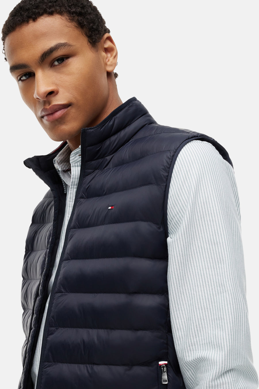 Bodywarmer bleu matelassé de TOMMY HILFIGER, avec petit logo drapeau et porté sur une chemise à rayures.