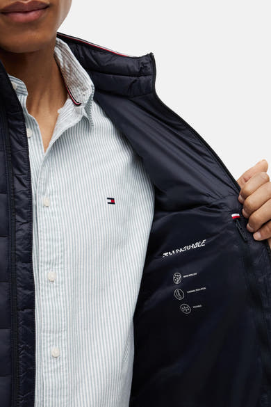 Gewatteerd blauw bodywarmer van TOMMY HILFIGER, open over een overhemd met fijne strepen, met binnenkant met prestatie-iconen en driekleurige kraag.