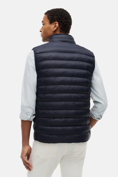 Blauwe bodywarmer van TOMMY HILFIGER, gewatteerd, van achteren gedragen over een gestreept overhemd en een crèmekleurige broek.