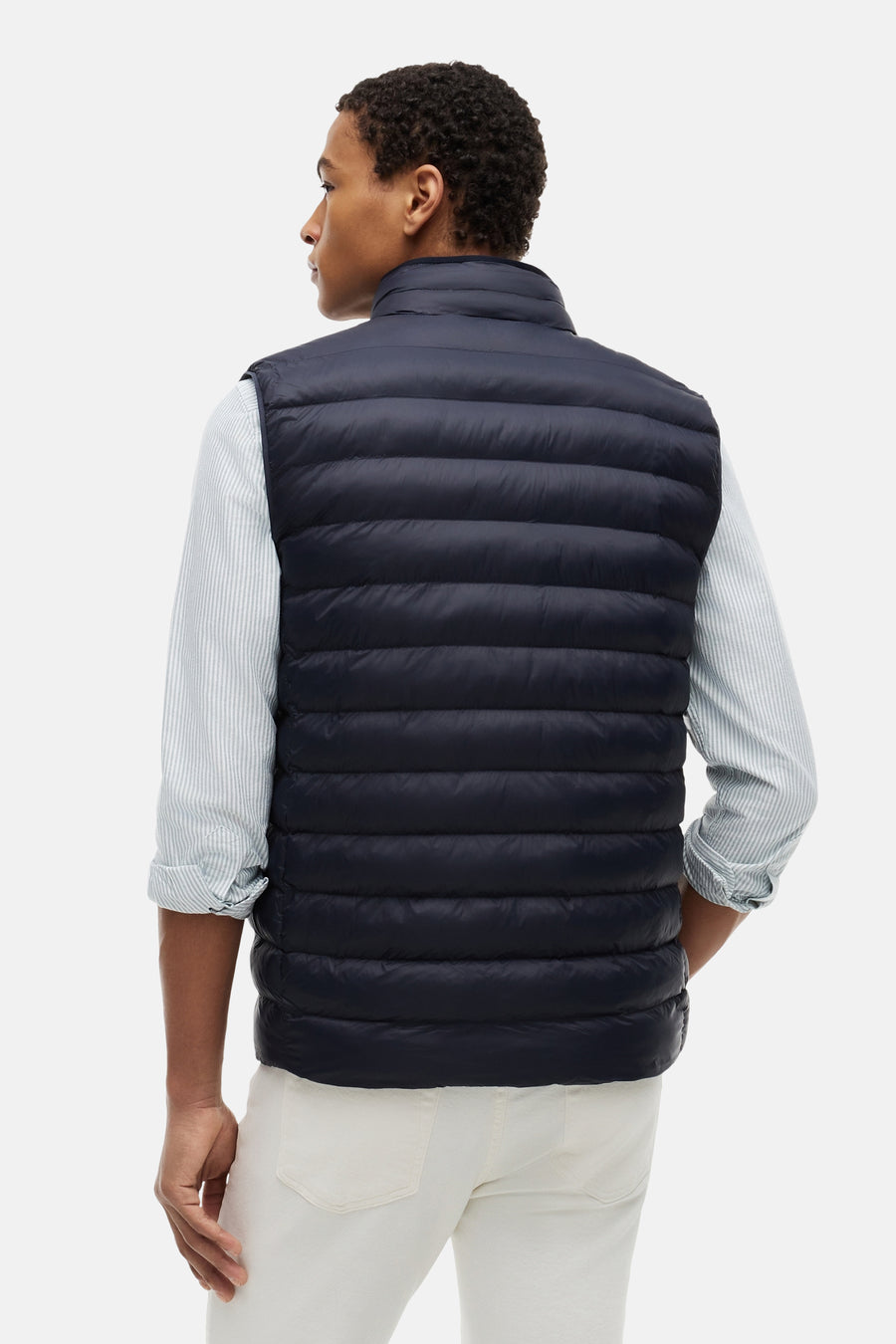 Blauwe bodywarmer van TOMMY HILFIGER, gewatteerd, van achteren gedragen over een gestreept overhemd en een crèmekleurige broek.