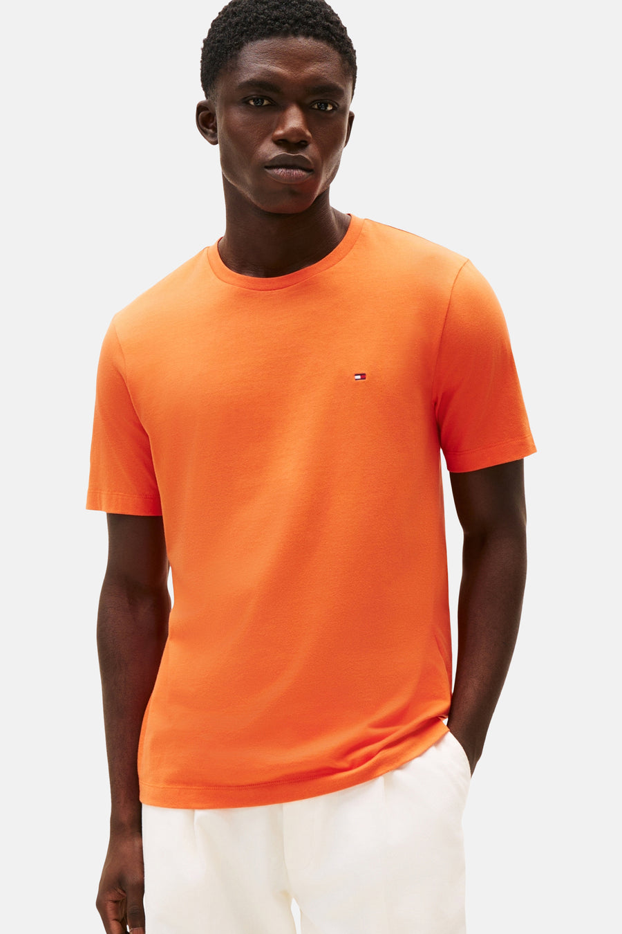 T-shirt orange à manches courtes de TOMMY HILFIGER, porté avec un pantalon blanc, sur fond clair.