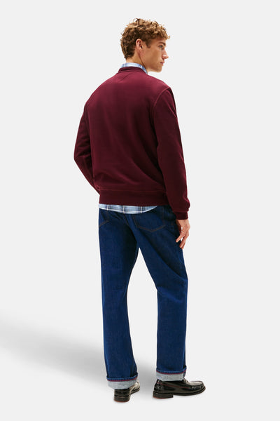 Bordeauxrode ronde hals sweatshirt van Tommy Hilfiger, gedragen over een geruit overhemd, met een spijkerbroek en zwarte loafers.