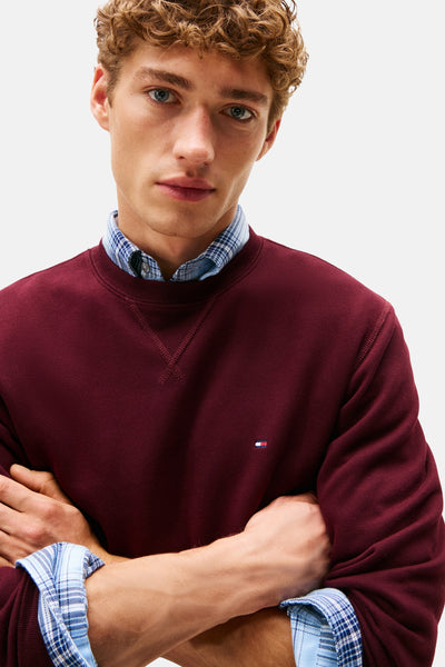 Sweat col rond rouge de Tommy Hilfiger, porté sur une chemise à carreaux, modèle croisé les bras.
