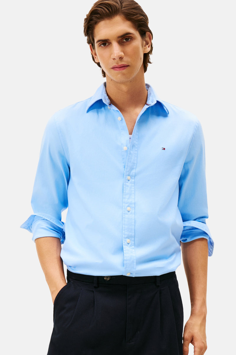 Chemise à manches longues bleue de TOMMY HILFIGER, portée avec un pantalon noir plissé, les manches retroussées.