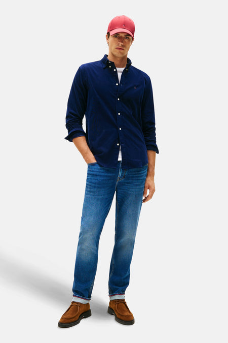Blauwe Tommy Hilfiger corduroy overhemd, open gedragen over een wit T-shirt, met een spijkerbroek en een rode pet.