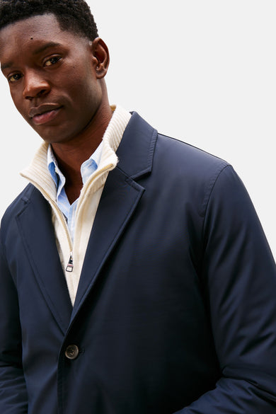 Lange blauwe jas van TOMMY HILFIGER, gedragen over een wollen trui met rits en een lichtblauw overhemd.