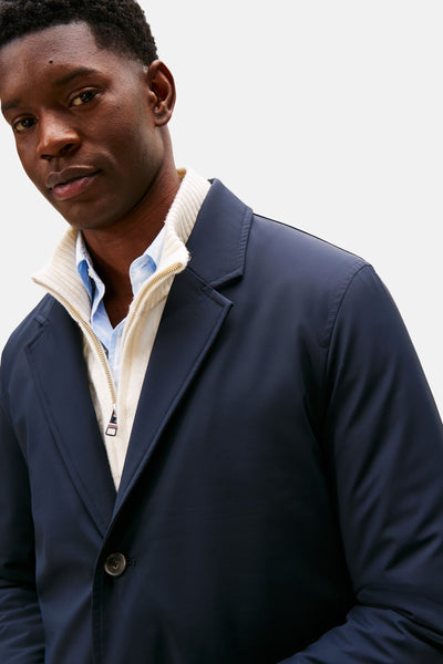 Lange blauwe jas van TOMMY HILFIGER, gedragen over een wollen trui met rits en een lichtblauw overhemd.