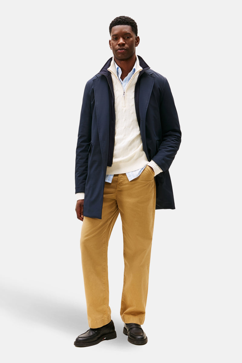 Lange blauwe jas van TOMMY HILFIGER, gedragen over een witte trui met rits en een bruine broek.