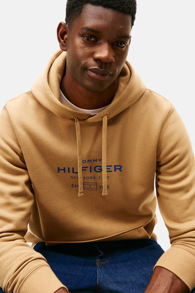 Beige hoodie van TOMMY HILFIGER, met marineblauw logo op de voorkant, gedragen met een blauwe jeans.
