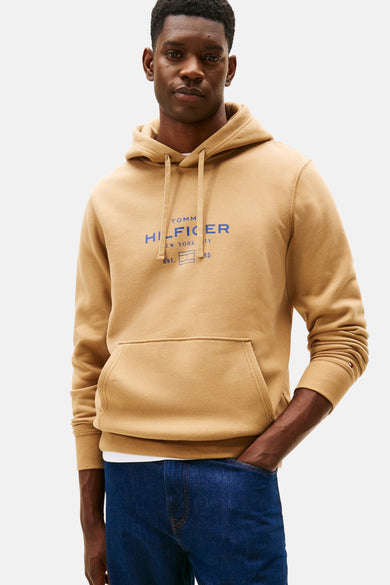 Beige hoodie van TOMMY HILFIGER, met blauw logo op de voorkant gedrukt, gedragen met een blauwe jeans.