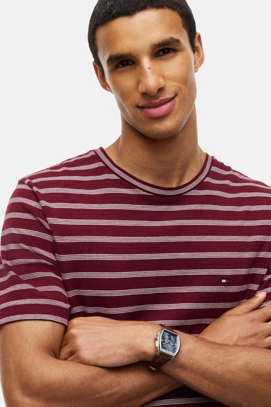 Red striped short-sleeve t-shirt by Tommy Hilfiger, featuring white horizontal stripes and small logo on chest, worn with brown leather watch, front view.