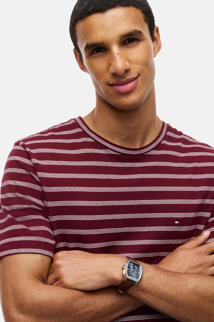 Red striped short-sleeve t-shirt by Tommy Hilfiger, featuring white horizontal stripes and small logo on chest, worn with brown leather watch, front view.