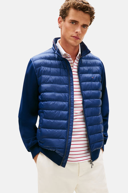 Blauwe jas van TOMMY HILFIGER, met contrasterende lange mouwen, gedragen over een gestreept rood poloshirt en een witte broek met bruine loafers.
