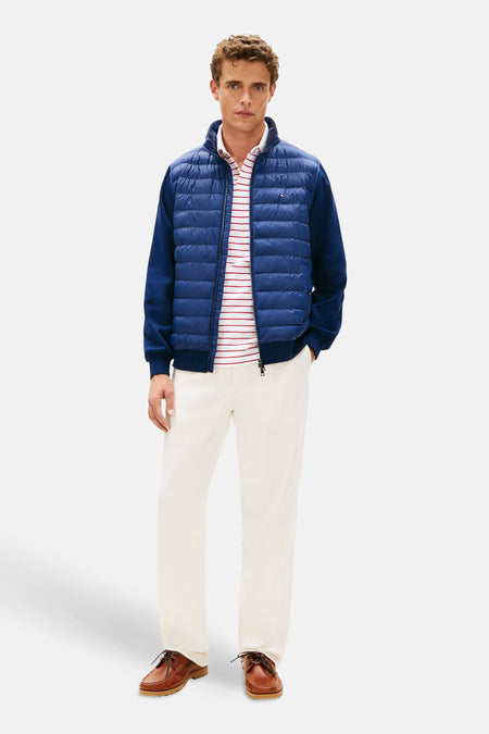 Veste bleu de TOMMY HILFIGER, avec manches longues contrastées, portée sur un polo rayé rouge et pantalon blanc et mocassins marrons.
