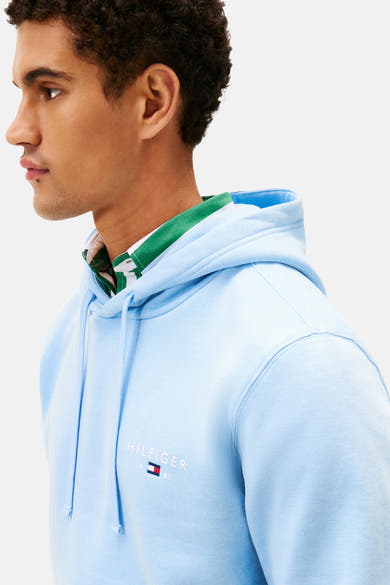 Blauwe Tommy Hilfiger hoodie met trekkoord en geborduurd logo op de borst, gedragen over een geruit overhemd.