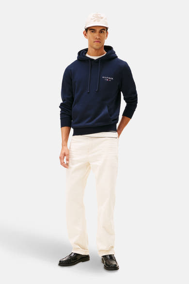 Sweat à capuche bleu de Tommy Hilfiger, porté avec un t-shirt blanc, un jean écru et des mocassins noirs, avec une casquette.