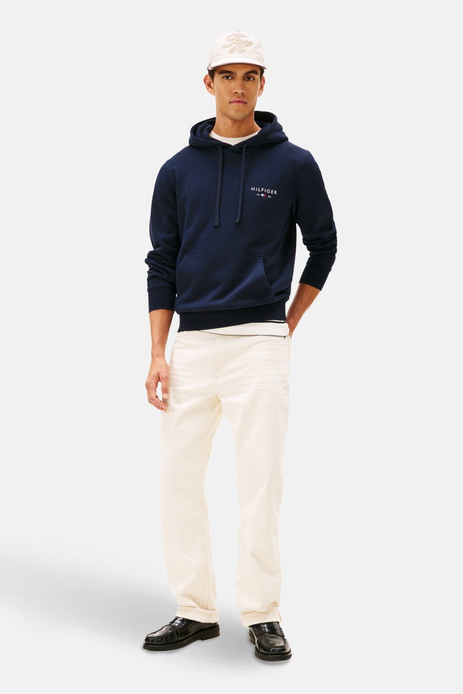 Sweat à capuche bleu de Tommy Hilfiger, porté avec un t-shirt blanc, un jean écru et des mocassins noirs, avec une casquette.