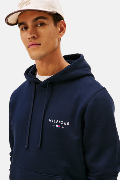 Sweat à capuche bleu de TOMMY HILFIGER avec cordon de serrage, logo sur le devant et casquette beige.
