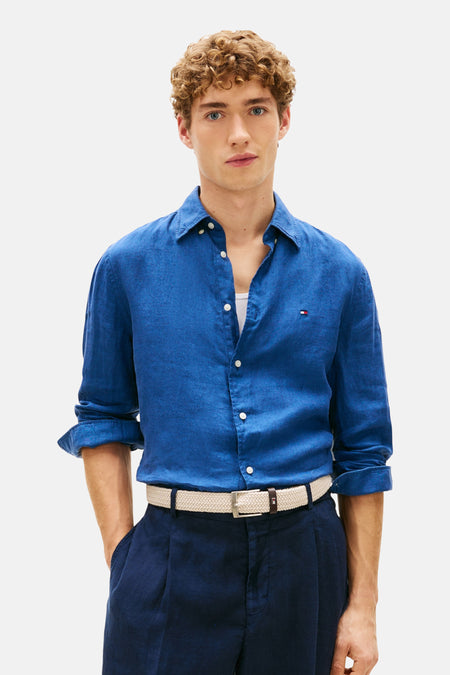 Chemise à manches longues bleue de Tommy Hilfiger, portée ouverte, avec pantalon bleu marine et ceinture en tissu tressé.