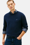 Pull à col rond bleu de TOMMY HILFIGER porté sur une chemise à carreaux, avec un jean.
