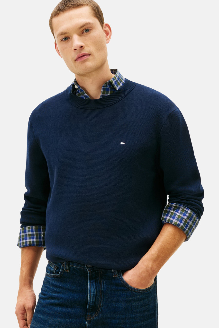 Pull à col rond bleu de TOMMY HILFIGER porté sur une chemise à carreaux, avec un jean.

