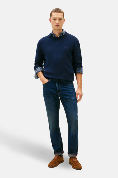 Pull à col rond bleu de TOMMY HILFIGER, porté sur une chemise à carreaux, avec un jean bleu et des chaussures marron.
