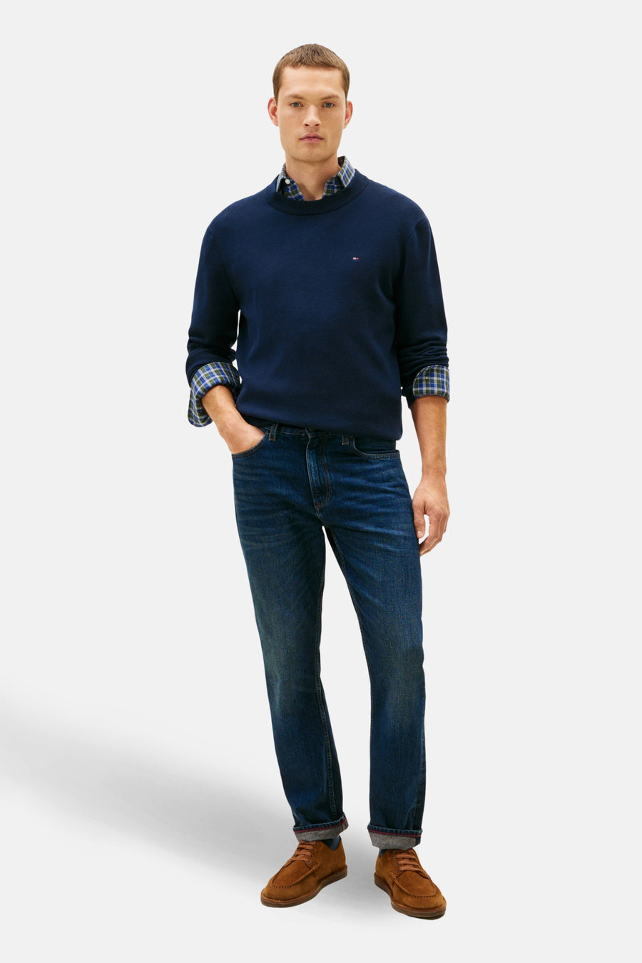 Pull à col rond bleu de TOMMY HILFIGER, porté sur une chemise à carreaux, avec un jean bleu et des chaussures marron.
