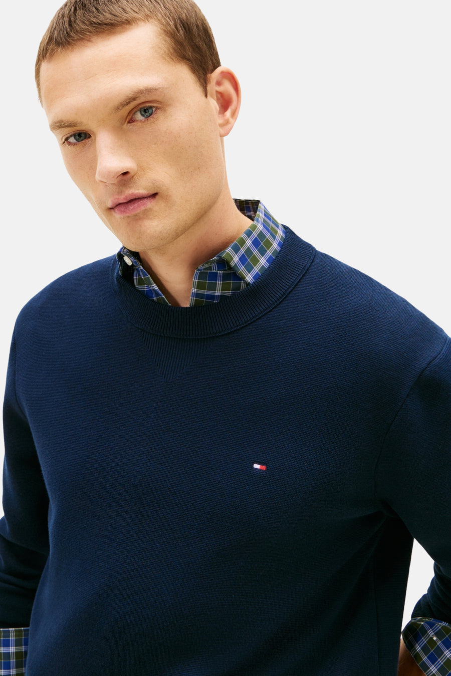 Pull à col rond bleu de TOMMY HILFIGER, porté sur une chemise à carreaux, logo sur la poitrine.