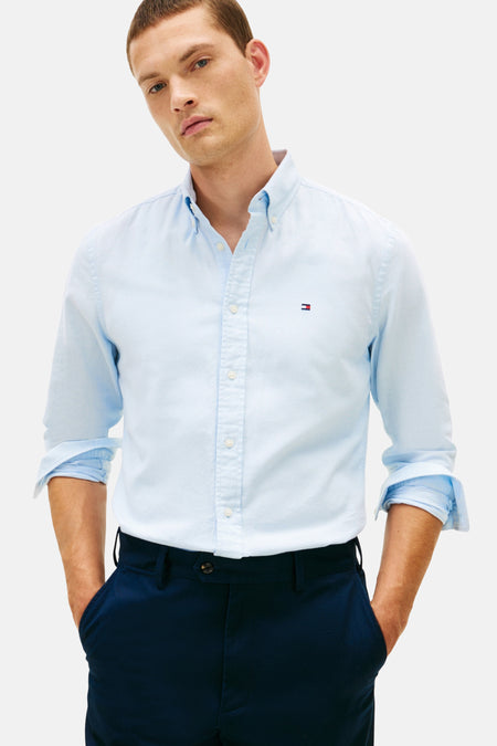 Chemise à manches longues bleue de TOMMY HILFIGER, avec manches retroussées et logo brodé, portée avec un pantalon bleu marine.