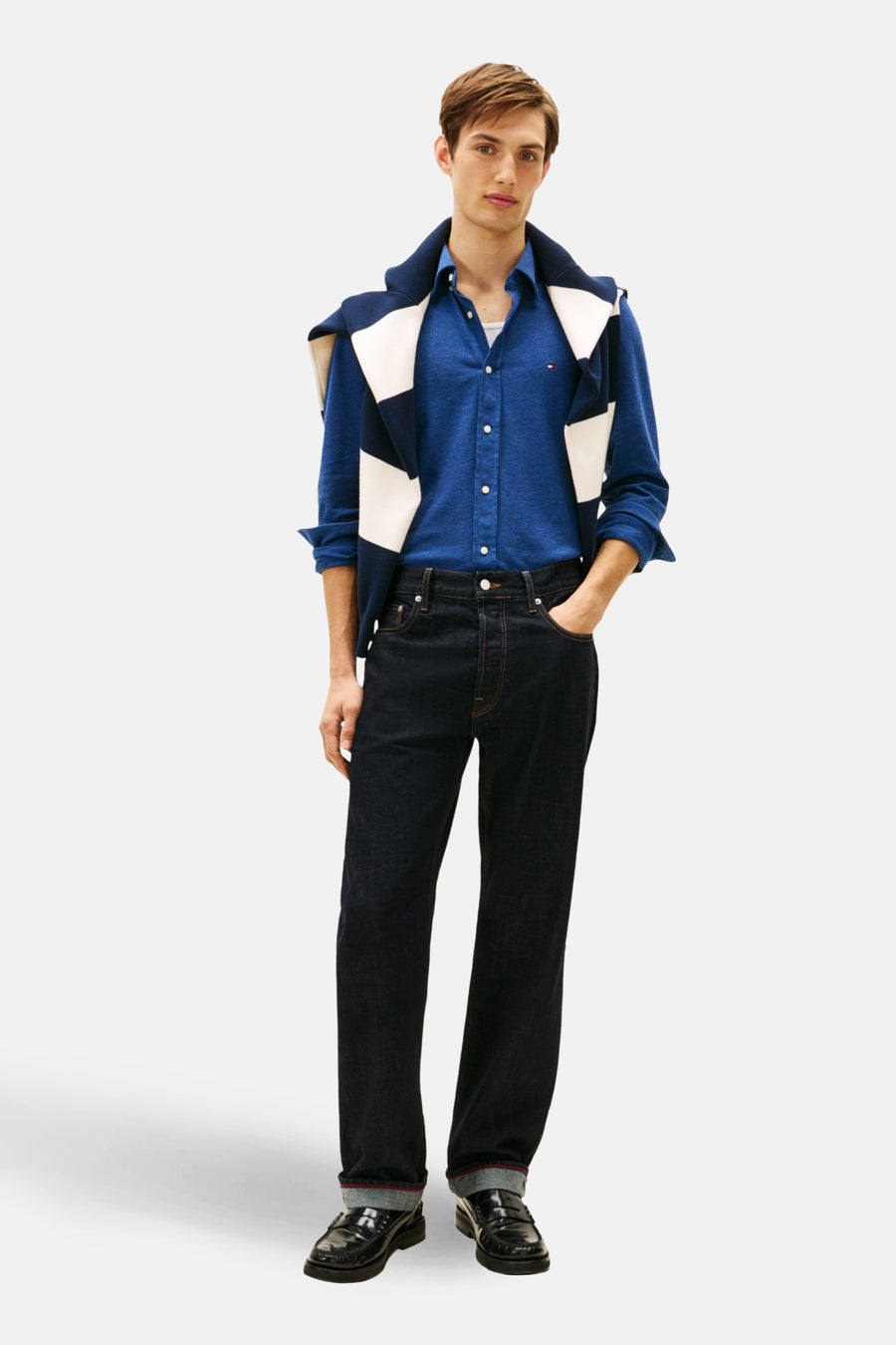 Chemise à manches longues bleue de TOMMY HILFIGER, portée avec un pull rayé et un jean foncé.
