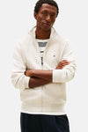 Gilet écru de Tommy Hilfiger, avec fermeture éclair et logo discret, porté sur un t-shirt rayé et un pantalon bleu marine.