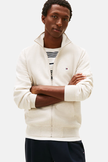 Gilet écru de Tommy Hilfiger, avec fermeture éclair et logo discret, porté sur un t-shirt rayé et un pantalon bleu marine.