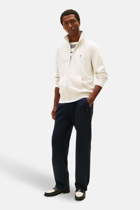 Gilet écru de TOMMY HILFIGER, porté sur un t-shirt rayé, avec un pantalon de survêtement bleu marine et des mocassins noirs et blancs.