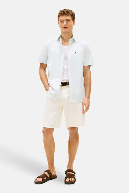 Chemise à manches courtes blanche à rayures par Tommy Hilfiger, portée ouverte sur un débardeur, avec un short beige et des sandales marron.
