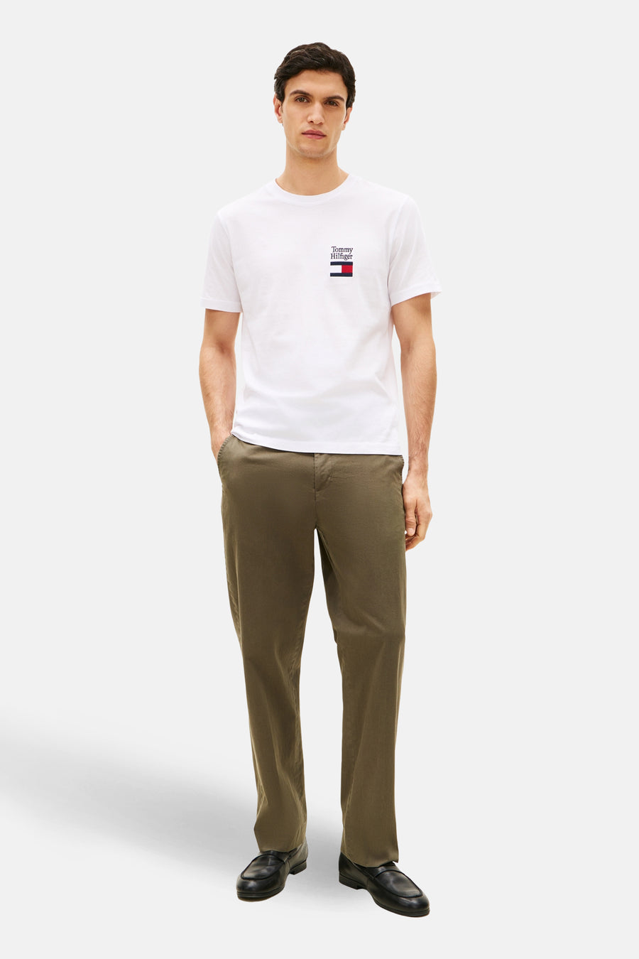 T-shirt blanc à manches courtes de TOMMY HILFIGER, avec logo brodé sur la poitrine, porté avec un pantalon et des chaussures noirs.