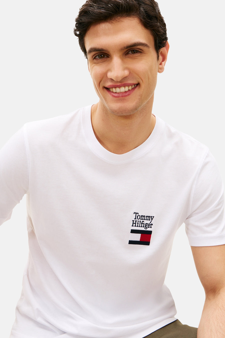 T-shirt blanc à manches courtes de Tommy Hilfiger, avec logo brodé sur la poitrine, porté par un homme souriant.
