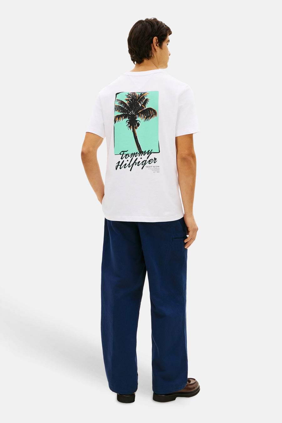 T-shirt blanc à manches courtes de TOMMY HILFIGER, vu de dos, avec un imprimé palmier graphique vert-bleu et noir, porté avec un pantalon bleu marine.