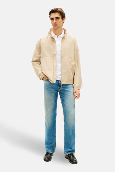 Veste beige de TOMMY HILFIGER, portée avec un polo blanc et un jean décontracté, sur fond uni.