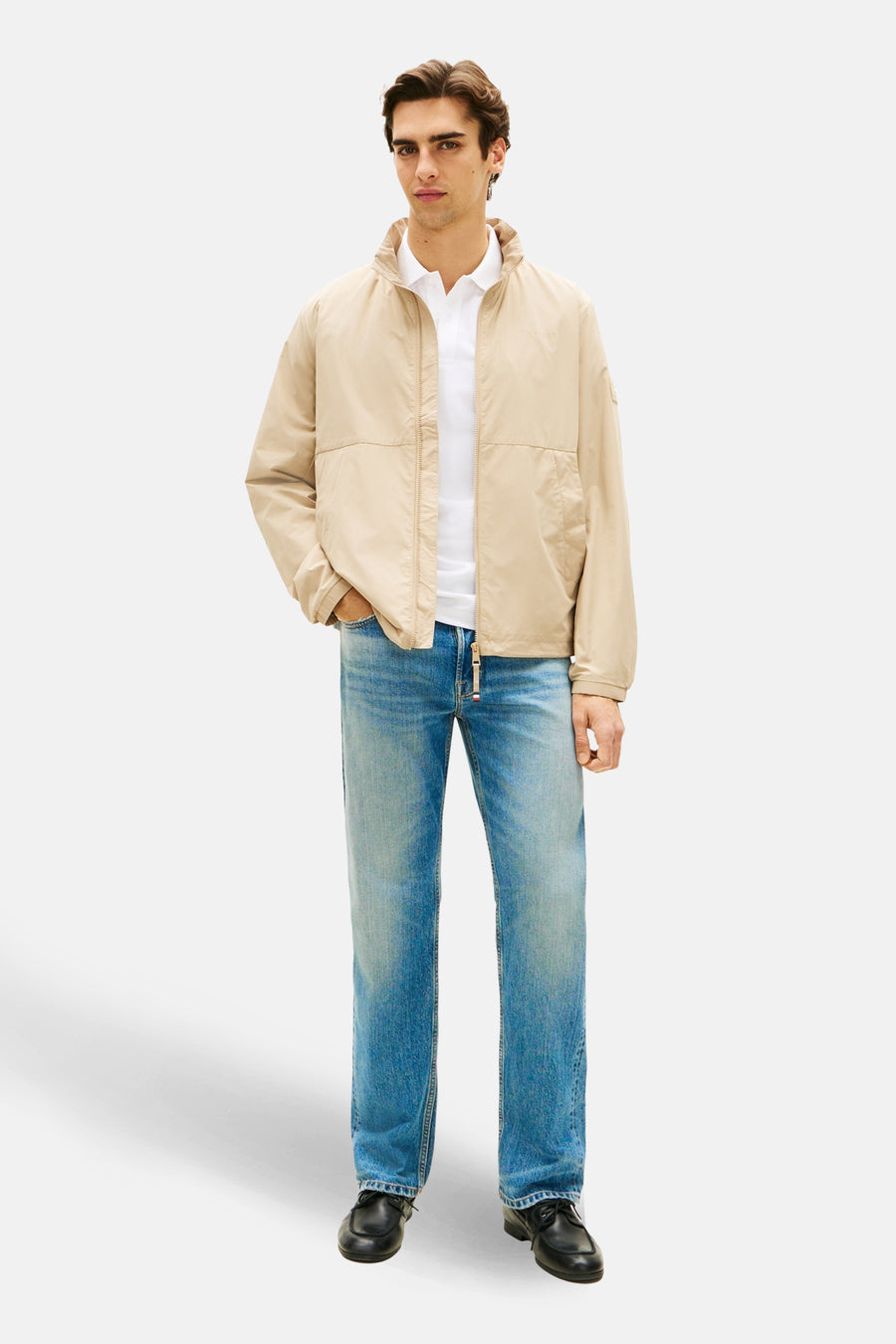 Veste beige de TOMMY HILFIGER, portée avec un polo blanc et un jean décontracté, sur fond uni.