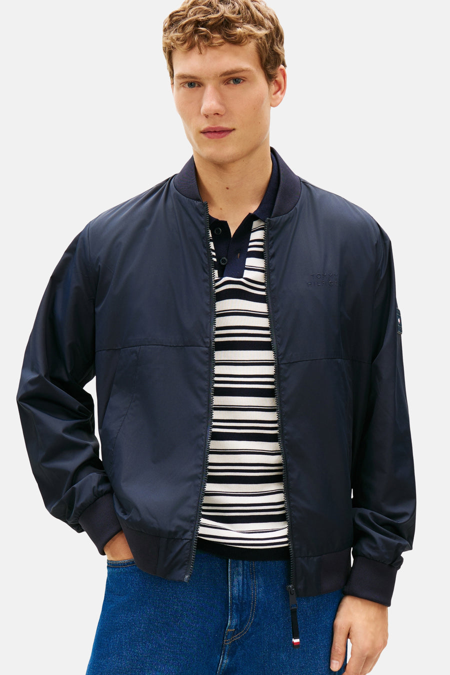 Veste bleu marine de TOMMY HILFIGER, zippée, portée sur un polo rayé, avec un jean.