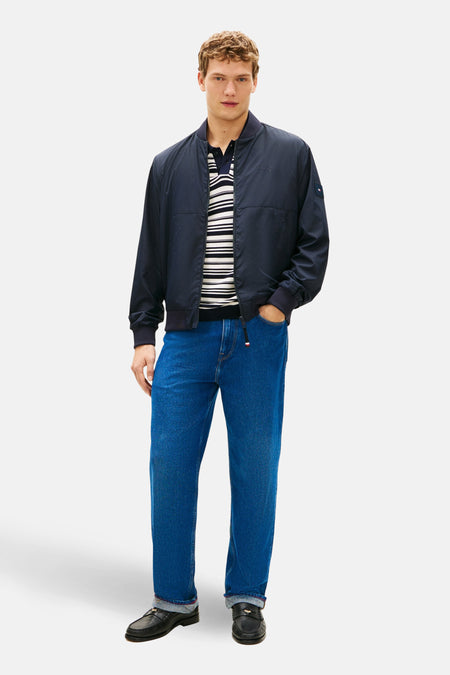 Veste bleu marine par Tommy Hilfiger, portée avec un polo rayé et un jean bleu, et des mocassins noirs.
